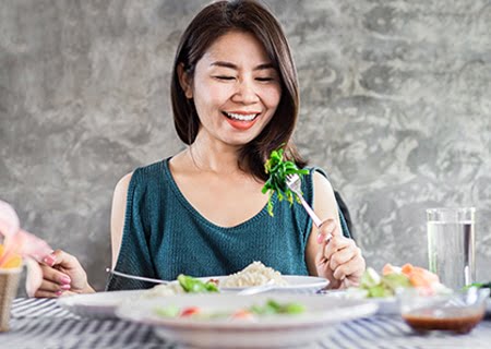 Mulher sentada à mesa segurando um garfo com folhas verdes de salada espetadas.