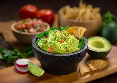 Mesa com guacamole em pote. Ao fundo, pedaços de frutas e chips