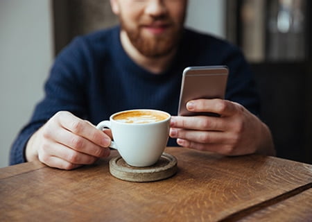 Homem segurando uma xícara de café com uma mão e, com a outra, olha o celular