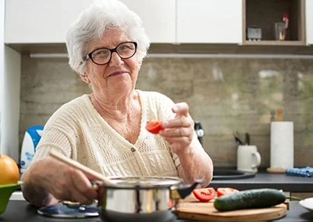Senhora sentada em bancada na cozinha comendo um tomate. Ela tem cabelos brancos curtos, usa óculos e sorri.