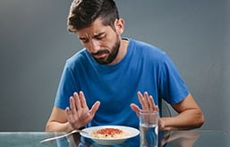 Homem sentado, na frente dele, sobre uma mesa, tem um copo d'água e um prato de macarrão com molho de tomate, mas ele olha para o alimento e gesticula com as duas mãos espalmadas sobre ele em um gesto de negação