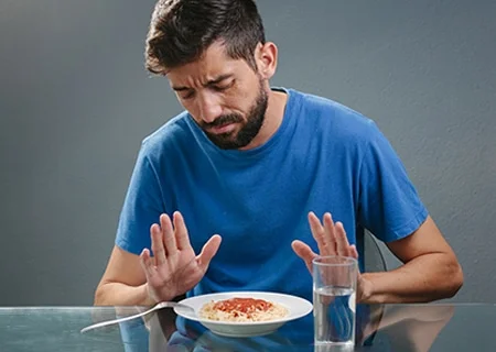 Homem sentado, na frente dele, sobre uma mesa, tem um copo d'água e um prato de macarrão com molho de tomate, mas ele olha para o alimento e gesticula com as duas mãos espalmadas sobre ele em um gesto de negação