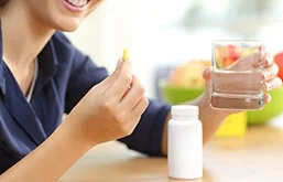 Mulher tomando vitamina. Ela está sentada em uma mesa e, com uma mão, segura uma pílula de vitamina e, na outra, um copo de água. Na mesa tem um potinho branco de vitamina. A mulher sorri.