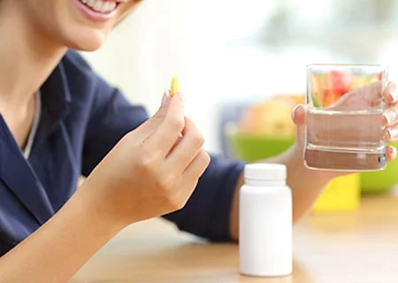 Mulher tomando vitamina. Ela está sentada em uma mesa e, com uma mão, segura uma pílula de vitamina e, na outra, um copo de água. Na mesa tem um potinho branco de vitamina. A mulher sorri.