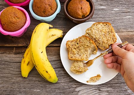 Mesa com muffins de banana, bananas e um prato com pedaços da receita e uma mão segurando um garfo o pegando