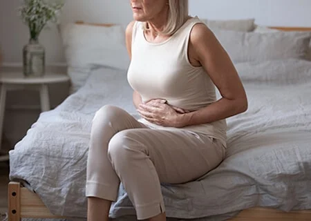 Mulher idosa sentada na cama com as mãos na barriga, sinalizando desconforto.