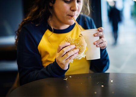 Menina sentada comendo hambúrguer e tomando refrigerante. <a href='https://br.freepik.com/fotos-vetores-gratis/alimento'>Alimento foto criado por rawpixel.com - br.freepik.com</a>