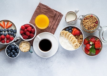 Mesa com tigelas com frutas, cereais, copo de suco, leite em jarra e café em xícara