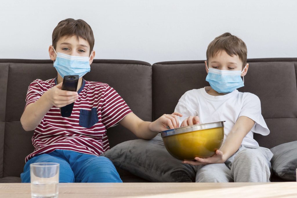 Garotinhos sentados em sofá com máscara e comendo pipoca em balde. Um segura controle da TV.