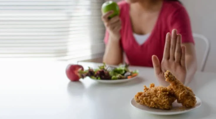 Mulher sentada à mesa comendo uma maçã verde. Na frente dela, um prato de salada. Mais à frente, um prato de carne frita, que ela recusa com a mão em sinal de recusa.