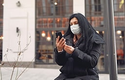 Mulher sentada com máscara olhando o celular. Imagem: Freepik