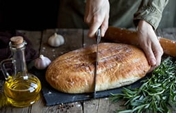 Pão sobre mesa ao lado de ramo de alecrim. Uma pessoa corta o pão ao meio.