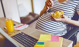 Mulher grávida sentada em frente a computador comendo laranja em pote