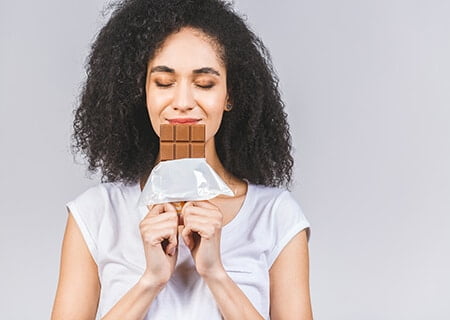Mulher segura barra de chocolate com as duas mãos em frente à boca. Ela tem os olhos fechados e sorri.