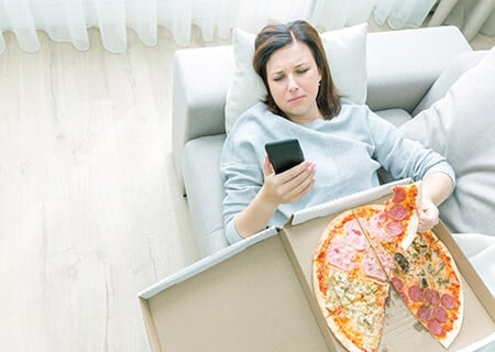 Mulher deitada no sofá com caixa de pizza sobre o colo e olhando o celular.