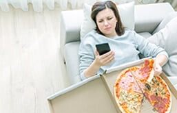 Mulher deitada no sofá com caixa de pizza sobre o colo e olhando o celular.