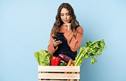 Mulher atrás de caixa de madeira com verduras olhando o celular. Ela está com a mão no queixo e expressão pensativa
