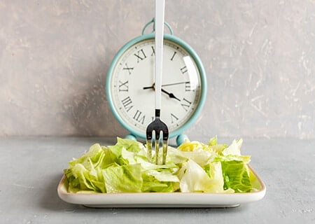 Prato de salada com garfo espetado e relógio de ponteiro ao fundo.