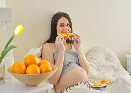 Mulher grávida sentada na cama comendo laranja