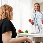 Mulher no consultório sentada olhando para nutricionista que, em pé, segura alimentos nas mãos