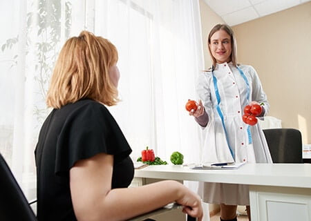 Mulher no consultório sentada olhando para nutricionista que, em pé, segura alimentos nas mãos