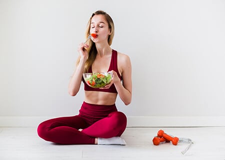 Mulher comendo salada sentada ao lado de halteres