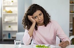 Mulher apática sentada na mesa com um prato de salada. Ela apoia uma mão na cabeça e tem expressão triste.