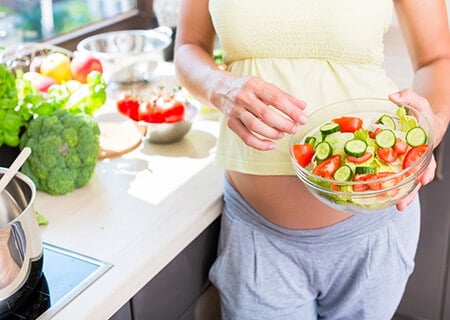 Mulher grávida na cozinha segurando pote com saladas