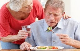 Homem comendo prato de salada sentado na mesa e mulher põe a mão em seu ombro e lhe oferece um copo de água