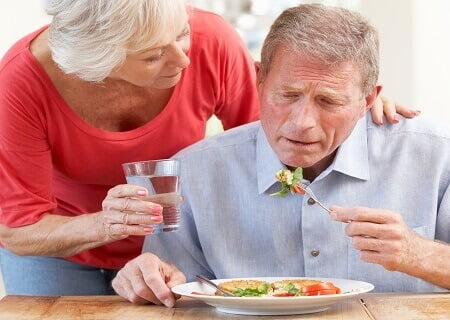 Homem comendo prato de salada sentado na mesa e mulher põe a mão em seu ombro e lhe oferece um copo de água
