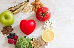 Alimentos bons para o coração em prato em forma de coração, entre frutas e grãos. Ao lado, estetoscópio e maçã verde, sobre mesa azul