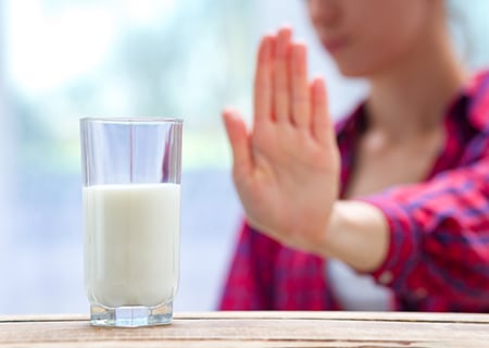 Copo de leite em cima da mesa e, ao fundo, mulher com a mão levantada em sinal de "não"