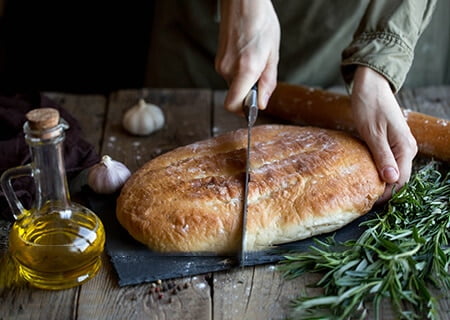 Pão sobre mesa ao lado de ramo de alecrim. Uma pessoa corta o pão ao meio.