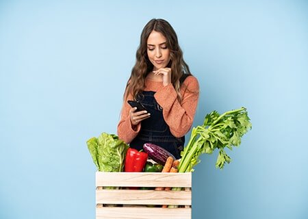 Mulher atrás de caixa de madeira com verduras olhando o celular. Ela está com a mão no queixo e expressão pensativa