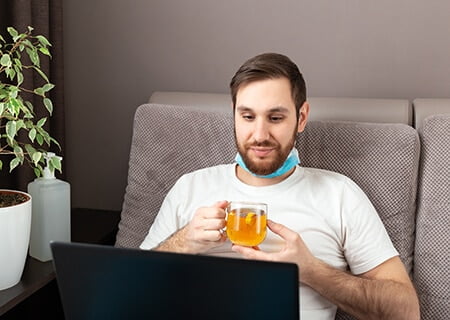 Homem sentado em sofá segurando xícara de chá com a máscara no queixo e olhando para o computador.