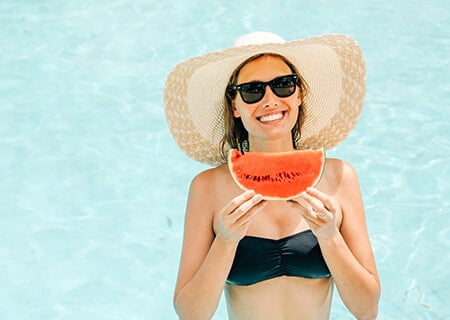 Mulher segurando fatia de melancia na piscina, um dos alimentos que ajudam a manter o bronzeado