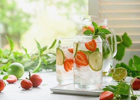 Mesa com bebidas em dois copos de vidro, com água, limão e morangos, que ajudam como hidratar o corpo de dentro para fora