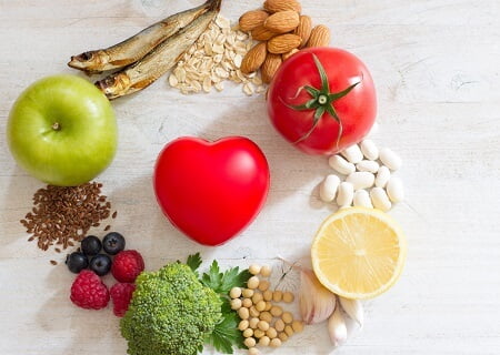 Alimentos bons para o coração em prato em forma de coração, entre frutas e grãos. Ao lado, estetoscópio e maçã verde, sobre mesa azul